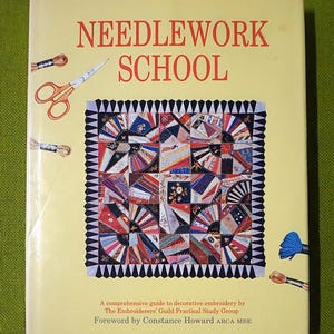 May include: A book titled "NEEDLEWORK SCHOOL" with red text on a light yellow background. The cover features a colorful patchwork design and illustrations of embroidery tools. The book is a comprehensive guide to decorative embroidery.