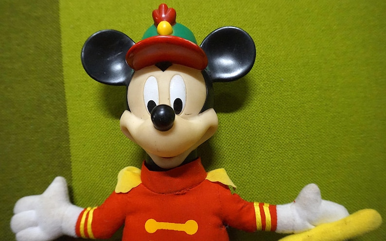Puede incluir: Una figura vintage de Mickey Mouse con uniforme de banda de m&uacute;sica. La figura tiene orejas negras, un sombrero rojo y una chaqueta roja con detalles amarillos. La figura sostiene una batuta amarilla y tiene guantes blancos.