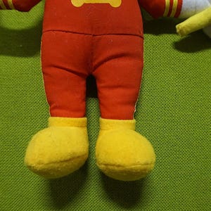 Puede incluir: Primer plano de las piernas y los pies de un peluche. El juguete tiene pantalones rojos y zapatos amarillos. El fondo es una tela verde texturizada.