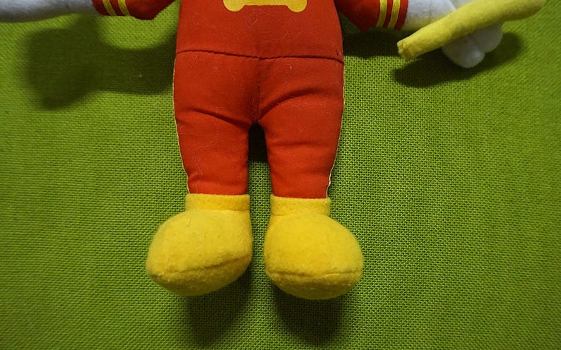 Puede incluir: Primer plano de las piernas y los pies de un peluche. El juguete tiene pantalones rojos y zapatos amarillos. El fondo es una tela verde texturizada.