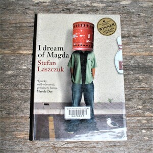 May include: A book cover with a red and white striped cylindrical object on the head of a person wearing a green shirt and blue jeans. The book title is "I dream of Magda" by Stefan Laszczuk. The book cover also includes a gold sticker that says "The Australian Vogel Winner Literary Award".