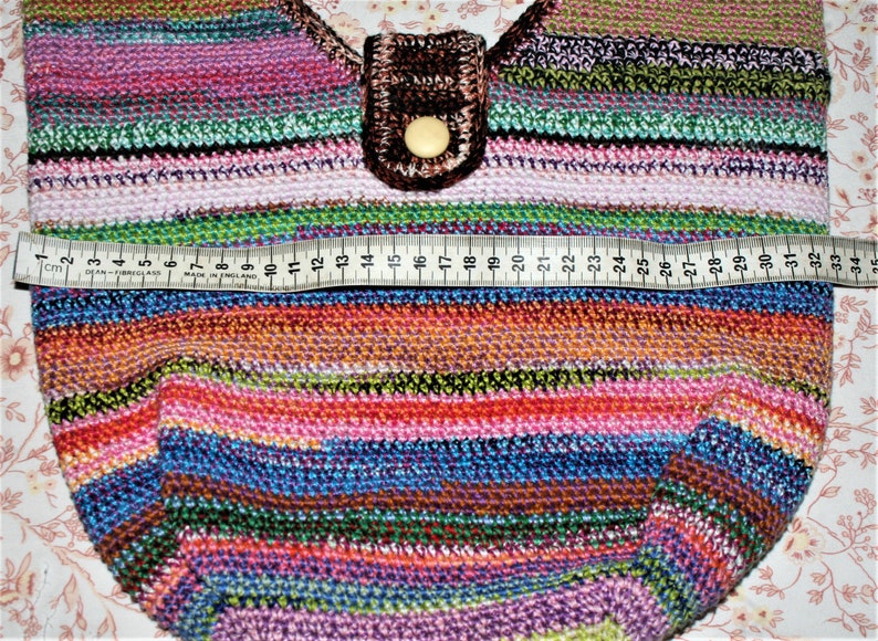 K&ouml;nnte beinhalten: Eine bunte gestreifte H&auml;keltasche mit einer braunen Lederklappe und einem Knopfverschluss. Die Tasche wird mit einem Ma&szlig;band gemessen, das eine Breite von 63,5 cm zeigt.