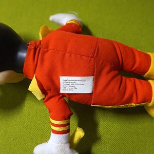 Puede incluir: Un peluche de un personaje de dibujos animados, con cabeza negra, guantes blancos y zapatos amarillos. El personaje viste un traje rojo con detalles amarillos. Una etiqueta blanca con texto est&aacute; cosida en la parte posterior del traje.