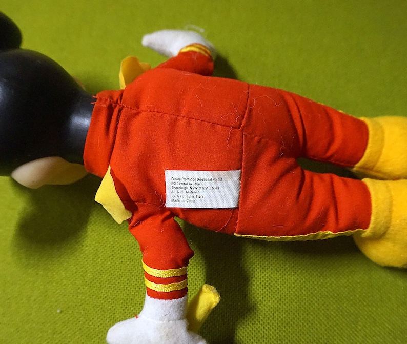 Puede incluir: Un peluche de un personaje de dibujos animados, con cabeza negra, guantes blancos y zapatos amarillos. El personaje viste un traje rojo con detalles amarillos. Una etiqueta blanca con texto est&aacute; cosida en la parte posterior del traje.
