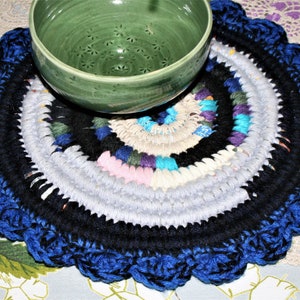 May include: A handmade, circular placemat with a scalloped blue edge. The center features concentric circles of black, gray, and colorful fabric scraps. A green bowl sits atop the placemat, suggesting a kitchen or dining theme.