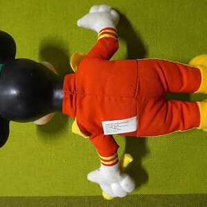 Puede incluir: Un peluche vintage de Mickey Mouse, visto desde arriba, tumbado sobre una superficie verde. El juguete presenta una cabeza negra con grandes orejas, un gorro verde y un mono rojo con detalles amarillos. Las manos y los pies son blancos y amarillos.