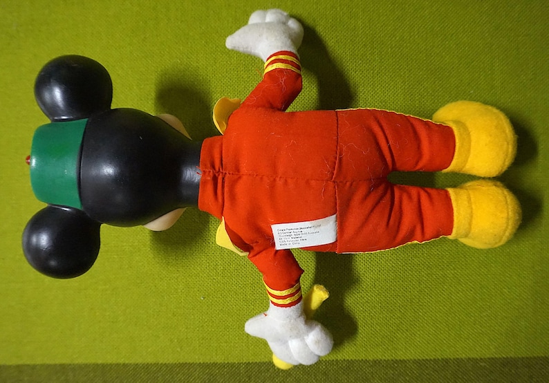 Puede incluir: Un peluche vintage de Mickey Mouse, visto desde arriba, tumbado sobre una superficie verde. El juguete presenta una cabeza negra con grandes orejas, un gorro verde y un mono rojo con detalles amarillos. Las manos y los pies son blancos y amarillos.