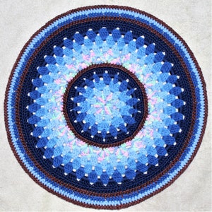 May include: A round crocheted mandala with a brown outer border and a center of blue, light blue, and pink. The mandala is made with a variety of stitches and has a circular design.