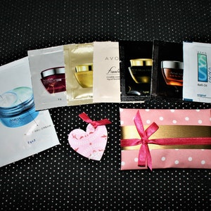 Puede incluir: Una caja de regalo rosa con lunares blancos y una cinta dorada atada con un lazo rosa. La caja está rodeada de pequeños productos de belleza en tamaño de muestra en paquetes de aluminio. Los paquetes muestran imágenes de cremas y lociones.