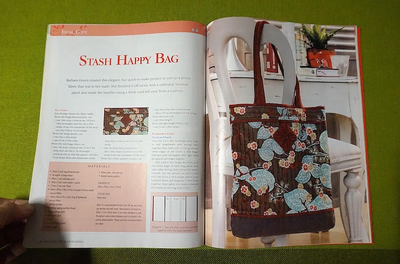 Puede incluir: Un bolso tote marr&oacute;n con un estampado floral de flores y hojas azules y rosas. El bolso tiene asas marrones oscuras y una base marr&oacute;n oscura. El bolso se muestra en una silla blanca junto a un libro abierto titulado "Stash Happy Bag".