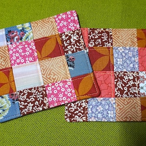 May include: Two square fabric coasters featuring a patchwork design of various cotton squares. The patterns include floral prints, geometric shapes, and solid colours in shades of pink, red, orange, blue, and white, all stitched together with visible seams.