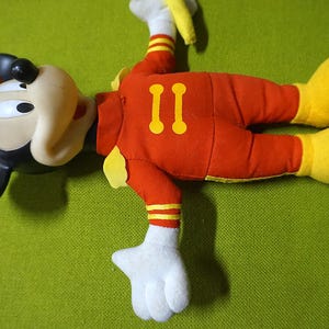 Puede incluir: Un peluche de Mickey Mouse con un traje de piloto. El juguete es principalmente rojo con detalles amarillos en los pies, guantes y el n&uacute;mero "II" en la espalda. El sombrero es rojo y verde, y las orejas son negras.