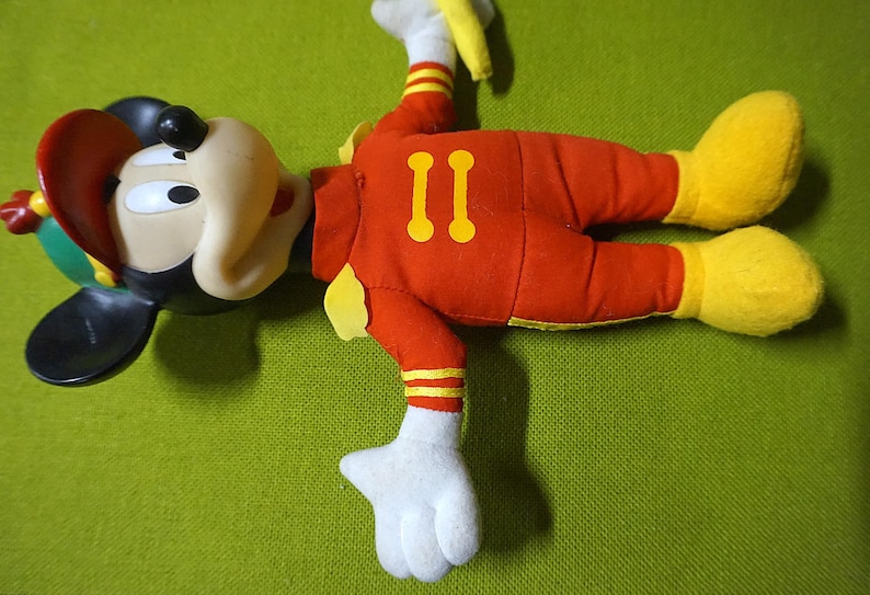 Puede incluir: Un peluche de Mickey Mouse con un traje de piloto. El juguete es principalmente rojo con detalles amarillos en los pies, guantes y el n&uacute;mero "II" en la espalda. El sombrero es rojo y verde, y las orejas son negras.
