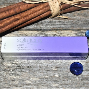 Puede incluir: Un tubo morado de crema de renovación para ojos Solutions Youth Minerals SPF 15, con una etiqueta blanca y el texto "Solutions Youth Minerals eye renewal cream SPF 15" en él. El tubo está sobre una superficie de madera con palitos de canela y cuentas de vidrio azules y verdes.