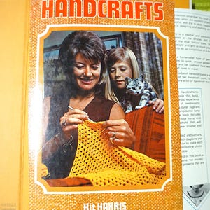 Könnte beinhalten: Ein Vintage-Buch mit dem Titel HANDCRAFTS von Kit Harris mit orangefarbenem Einband und einem zentralen Foto eines Erwachsenen und eines Kindes, die eine gelbe Decke häkeln, mit einer kleinen Katze auf einem braunen Sofa vor strukturiertem Hintergrund.