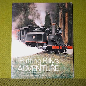 Pode incluir: Capa de livro com uma locomotiva a vapor preta, emitindo fumo branco, viajando por uma floresta. O título "Puffing Billy's ADVENTURE" é visível. Fundo verde.