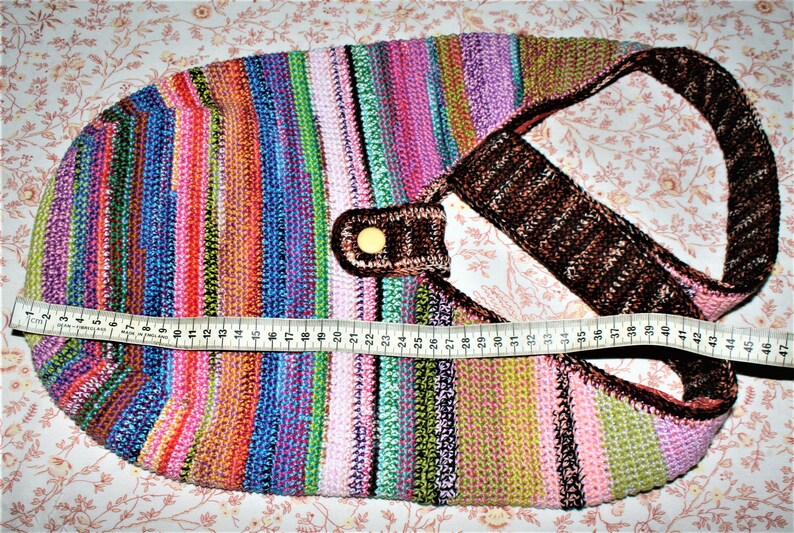 K&ouml;nnte beinhalten: Eine bunte gestreifte geh&auml;kelte Tragetasche mit einem braunen Ledergriff und einem goldenen Knopf. Die Tasche ist 119 cm lang.