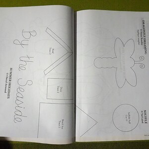 Puede incluir: Libro abierto con p&aacute;ginas que presentan dibujos lineales de una lib&eacute;lula, formas de casa y el texto "By the Seaside" y "Summer Holidays". Las p&aacute;ginas est&aacute;n sobre una superficie verde.