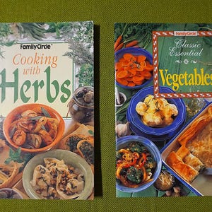 以下が含まれることがあります： 「Family Circle Cooking with Herbs」と「Family Circle Classic Essential Vegetables」という2冊の料理本が表示されています。表紙には、調理された料理、ハーブ、野菜の写真が掲載されています。本は緑色の背景に置かれています。