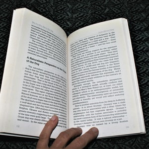 May include: Open book with a white background and black text. The book is open to a page titled "A Naturalistic Perspective on the Origins of the Dog". The text discusses the origins of the dog and its relationship with humans. A hand is holding the book open.