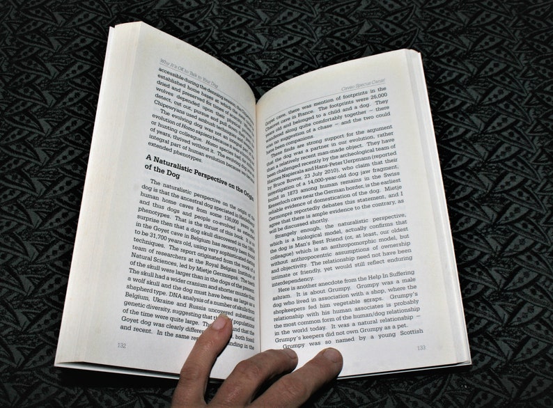 May include: Open book with a white background and black text. The book is open to a page titled "A Naturalistic Perspective on the Origins of the Dog". The text discusses the origins of the dog and its relationship with humans. A hand is holding the book open.