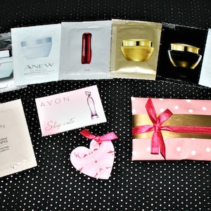 Puede incluir: Una caja de regalo rosa y blanca con lunares y una cinta dorada y rosa. La caja está rodeada de pequeños productos de belleza de muestra, incluyendo crema facial, suero y base de maquillaje. Los productos son de la marca Avon.