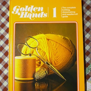 Könnte beinhalten: Ein Buch mit dem Titel "Golden Hands 1" mit gelbem Einband, das einen Wollknäuel, eine Schere, eine Garnrolle und einen Fingerhut zeigt. Das Buch ist ein umfassender Leitfaden für Stricken, Nähen und Handarbeiten.