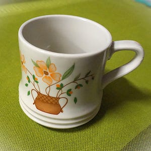 May include: A white ceramic mug with a curved handle. The mug features a floral design with orange flowers and green leaves in a brown basket. The mug has a ridged base.