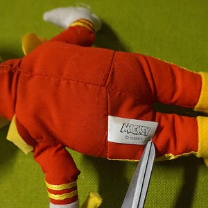 Puede incluir: Un peluche de Mickey Mouse, tumbado boca arriba, con un traje rojo y zapatos amarillos. El juguete tiene la cabeza negra y guantes blancos. Una etiqueta con la palabra "Mickey" es visible. Unas tijeras cortan la etiqueta.