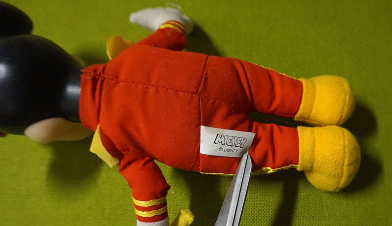 Puede incluir: Un peluche de Mickey Mouse, tumbado boca arriba, con un traje rojo y zapatos amarillos. El juguete tiene la cabeza negra y guantes blancos. Una etiqueta con la palabra "Mickey" es visible. Unas tijeras cortan la etiqueta.