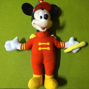Puede incluir: Un juguete de peluche vintage de Mickey Mouse con un uniforme rojo de banda de m&uacute;sica con detalles amarillos. El juguete tiene orejas negras, un sombrero rojo y guantes blancos, sosteniendo una batuta amarilla. El fondo es verde liso.