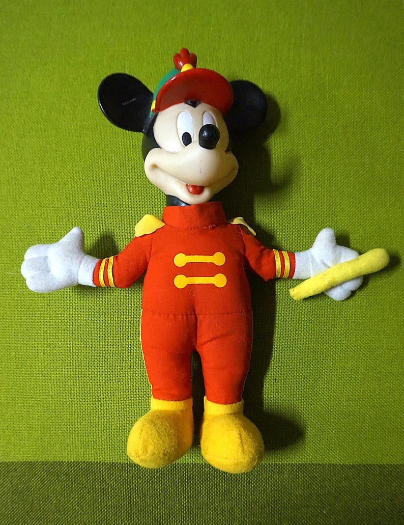 Puede incluir: Un juguete de peluche vintage de Mickey Mouse con un uniforme rojo de banda de m&uacute;sica con detalles amarillos. El juguete tiene orejas negras, un sombrero rojo y guantes blancos, sosteniendo una batuta amarilla. El fondo es verde liso.