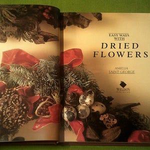 Puede incluir: Un libro titulado "Easy Ways with Dried Flowers" de Amelia Saint George, publicado por Weldon Publishing. La portada presenta un arreglo floral con cinta roja, ramas de pino y bayas secas.