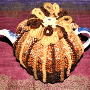 May include: A crocheted teapot cozy in brown, beige, and white with a decorative flower design on top. The cozy is on a teapot with a blue and white pattern.