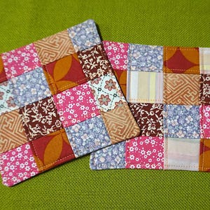 May include: Two square fabric coasters featuring a patchwork design composed of various small squares. The patterns include pink floral prints, light blue blossoms, geometric tan shapes, and dark red floral motifs, all stitched together with visible seams.