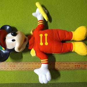 Puede incluir: Un peluche vintage de Mickey Mouse con uniforme rojo con detalles amarillos y el n&uacute;mero "11". El juguete tiene orejas negras, un sombrero rojo y verde y zapatos amarillos. Una regla de madera est&aacute; al lado del juguete.