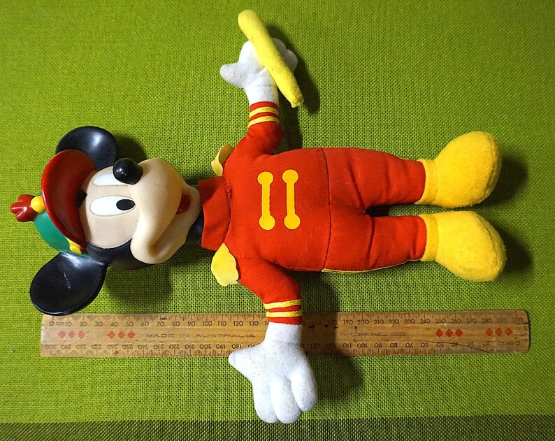 Puede incluir: Un peluche vintage de Mickey Mouse con uniforme rojo con detalles amarillos y el n&uacute;mero "11". El juguete tiene orejas negras, un sombrero rojo y verde y zapatos amarillos. Una regla de madera est&aacute; al lado del juguete.