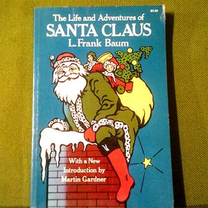 May include: A vintage paperback book cover featuring a cartoon illustration of Santa Claus climbing down a chimney. The book title is "The Life and Adventures of Santa Claus" by L. Frank Baum. The cover is blue with a yellow star and white icicles.