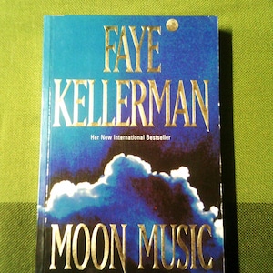 Könnte beinhalten: Ein Buchcover mit blauem und schwarzem Hintergrund, auf dem eine Wolkenformation zu sehen ist. Der Titel "MOON MUSIC" ist in goldenen Buchstaben geschrieben. Der Name des Autors "FAYE KELLERMAN" ist ebenfalls in goldenen Buchstaben geschrieben. Der Text "Her New International Bestseller" ist in weißen Buchstaben geschrieben. Der Text "A mesmerizing vision of America's dark and wounded heart" ist in weißen Buchstaben geschrieben.