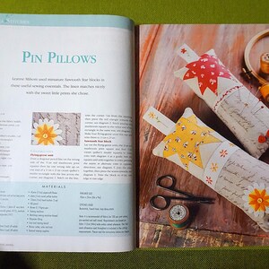 Puede incluir: Libro abierto con el t&iacute;tulo "Pin Pillows" y dos cojines decorativos para alfileres. Uno tiene un dise&ntilde;o de estrella roja, el otro un dise&ntilde;o de estrella amarilla. Tambi&eacute;n se ven unas tijeras y una bobina de hilo.