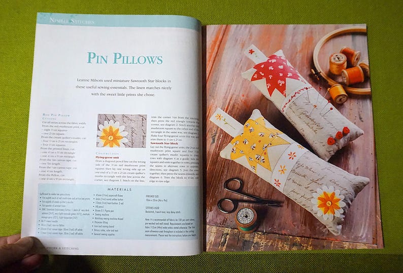 Puede incluir: Libro abierto con el t&iacute;tulo "Pin Pillows" y dos cojines decorativos para alfileres. Uno tiene un dise&ntilde;o de estrella roja, el otro un dise&ntilde;o de estrella amarilla. Tambi&eacute;n se ven unas tijeras y una bobina de hilo.