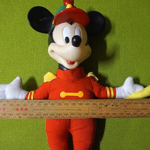 Puede incluir: Mu&ntilde;eco vintage de Mickey Mouse con uniforme rojo, detalles amarillos y gorro a juego. El mu&ntilde;eco tiene orejas negras, cara blanca y guantes blancos. Una regla de madera, de unos 25 cm, se sostiene sobre los brazos extendidos del mu&ntilde;eco.