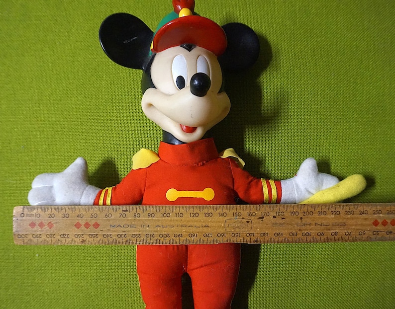 Puede incluir: Mu&ntilde;eco vintage de Mickey Mouse con uniforme rojo, detalles amarillos y gorro a juego. El mu&ntilde;eco tiene orejas negras, cara blanca y guantes blancos. Una regla de madera, de unos 25 cm, se sostiene sobre los brazos extendidos del mu&ntilde;eco.