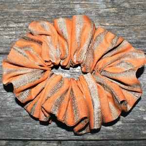 May include: Orange and brown striped fabric hair scrunchie. The scrunchie is made of a soft, stretchy fabric and is ideal for holding hair in place.