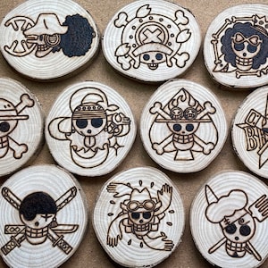 Pode incluir: Conjunto de 10  tábuas de madeira com diferentes desenhos de caveiras e ossos cruzados da popular série de anime One Piece. Cada tábua é circular e apresenta um design único com o símbolo de um personagem diferente.