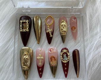May include: Set of ten burgundy and gold press-on nails with religious and angel themed designs. The nails feature gold chains, crosses, and images of the Virgin Mary and angels.