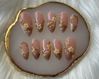 May include: A set of ten pink oval-shaped false nails with gold accents and white flowers. The nails are arranged on a white and gold tray.