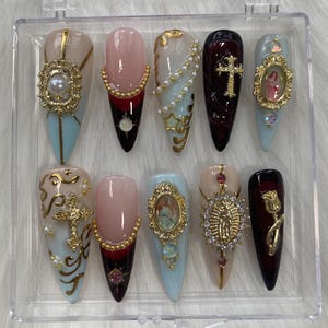 Può includere: Set di dieci unghie finte lunghe e appuntite con vari disegni a tema religioso. Le unghie sono dipinte in tonalità di blu, rosa e bordeaux. Ogni unghia presenta un accento dorato con una perla, una croce o una figura religiosa.
