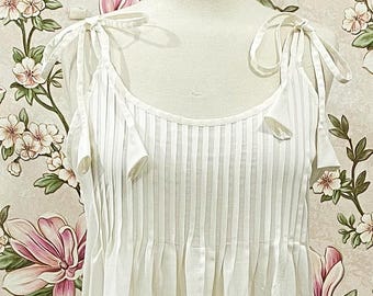May include: A white, floor-length dress with a pleated bodice and tie straps. The dress features a delicate embroidered hemline. The background is a floral wallpaper with pink and white flowers and green leaves.
