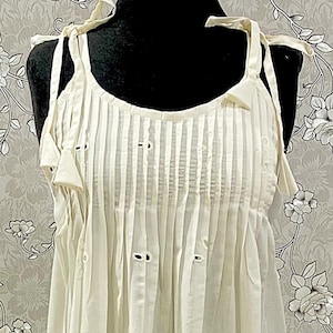 May include: A cream-coloured sleeveless dress featuring a round neckline with tie-up shoulder straps, vertical pleats across the chest, and an intricate eyelet embroidery pattern throughout the fabric, ending in a scalloped hemline detail.